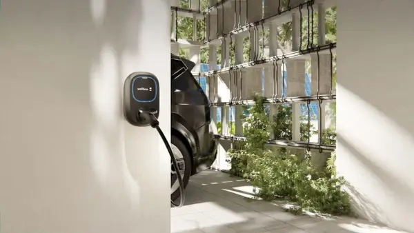 Installazione wallbox per auto elettrica, con cavo collegato a un veicolo scuro, in un ambiente moderno con vegetazione e pannelli solari.